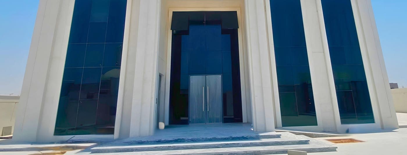 Front of a white stone villa with tall tinted glass windows and a glazed entrance